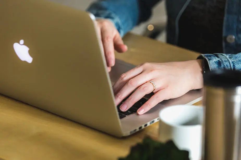 Nahaufnahme von Händen beim Tippen auf einer Laptop-Tastatur an einem Holztisch, im Vordergrund eine Kaffeetasse und ein Thermobecher.