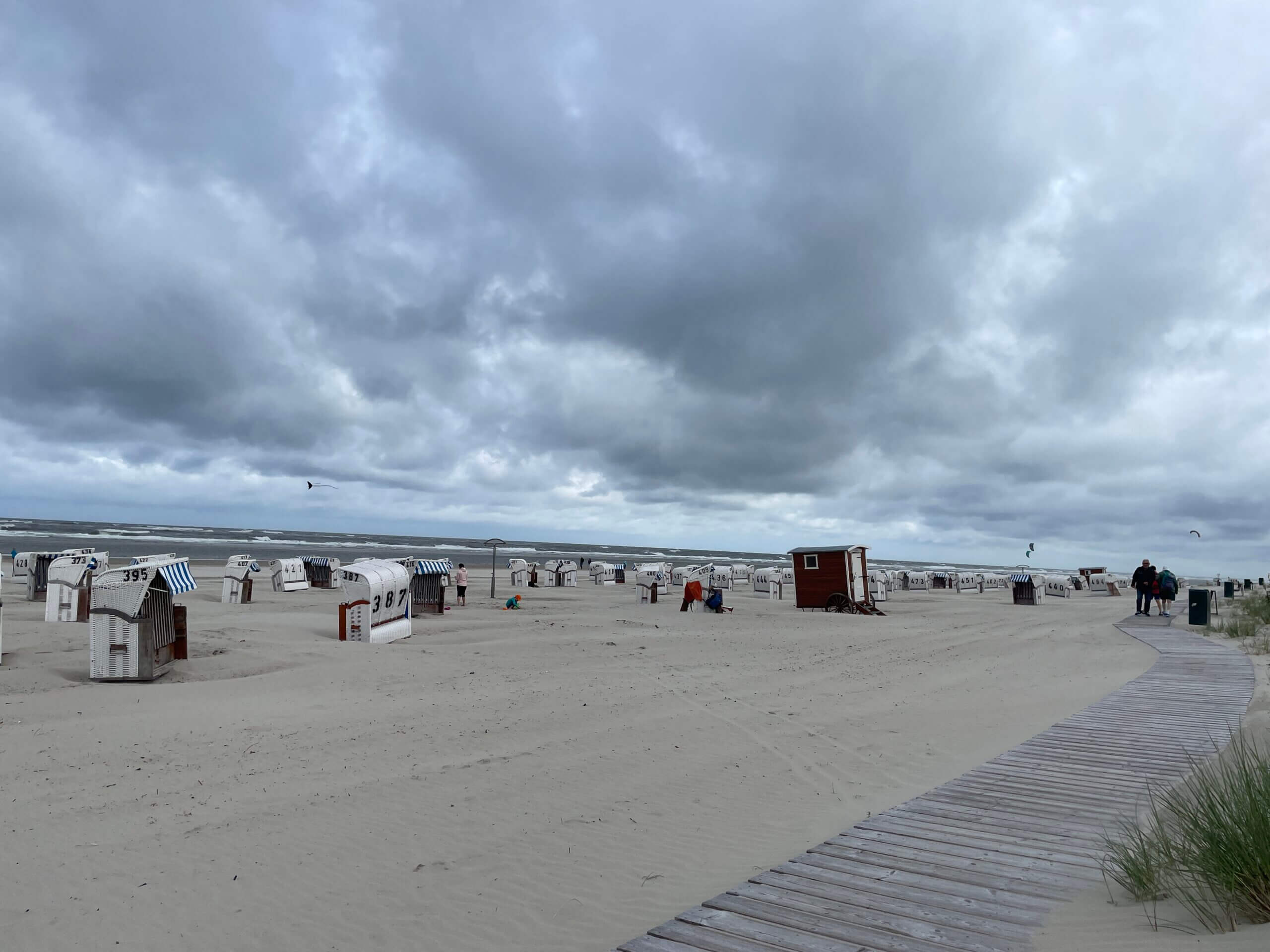 Typischer Spiekeroog-Strand mit weiß-blauen Strandkörben, Holzsteg und bewölktem Nordsee-Himmel.