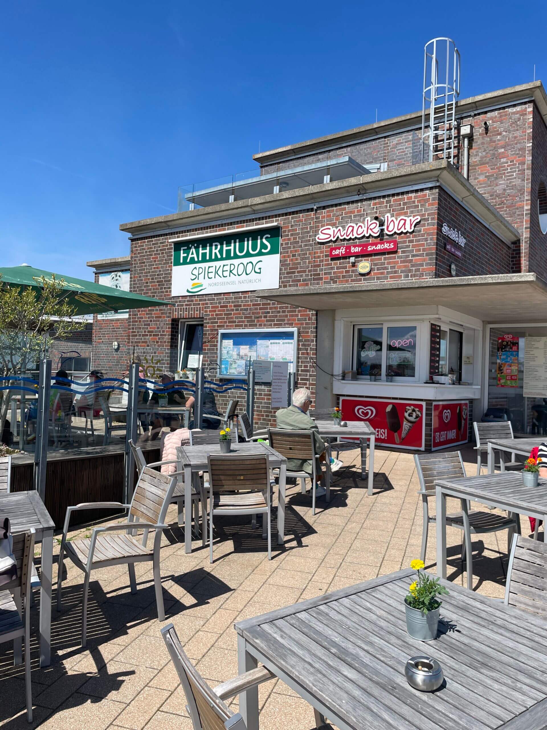 Fahrhaus Spiekeroog in Neuharlingersiel: Café und Snackbar mit Außenbereich, Holzmöbeln und blauem Himmel.